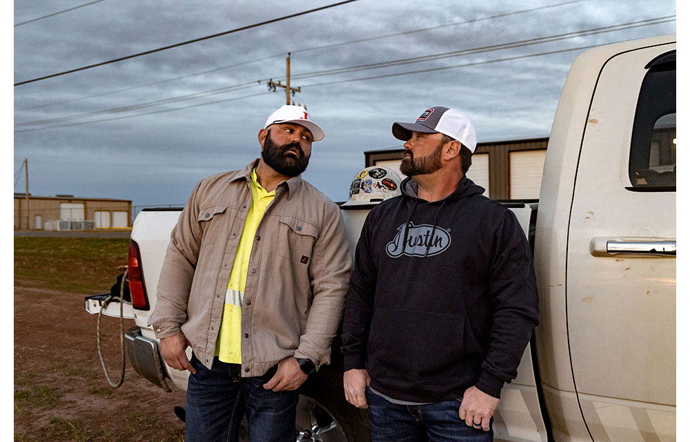 Two linemen, Kevin Walker and Dylan Edgmon, in workwear and ball caps stand beside a truck at dusk, talking casually near a job site with buildings and power lines in the background.